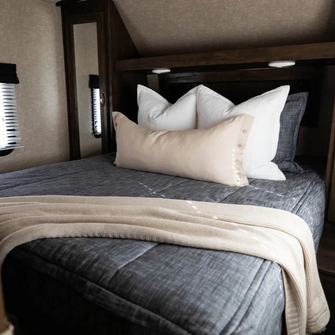 A neatly made bed features the Airstream RV Fit Beddy’s for Excella Travel Trailers in gray, complemented by three white pillows, a long beige pillow, and a beige knit throw blanket at the foot. Perfectly tailored RV mattress sheets ensure ultimate comfort. Beside the bed is a mirrored wardrobe, and a window with blinds is on the left.