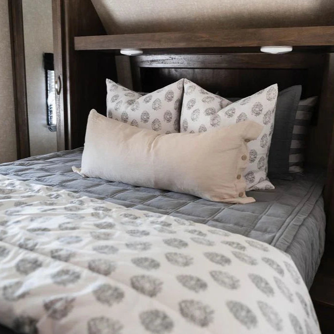 A neatly made bed featuring Airstream RV Fit Beddy’s for Flying Cloud Travel Trailers in gray with zippered bedding, paired with patterned white pillows, a long beige cushion, and a white duvet adorned with gray designs, all set against a wooden headboard.