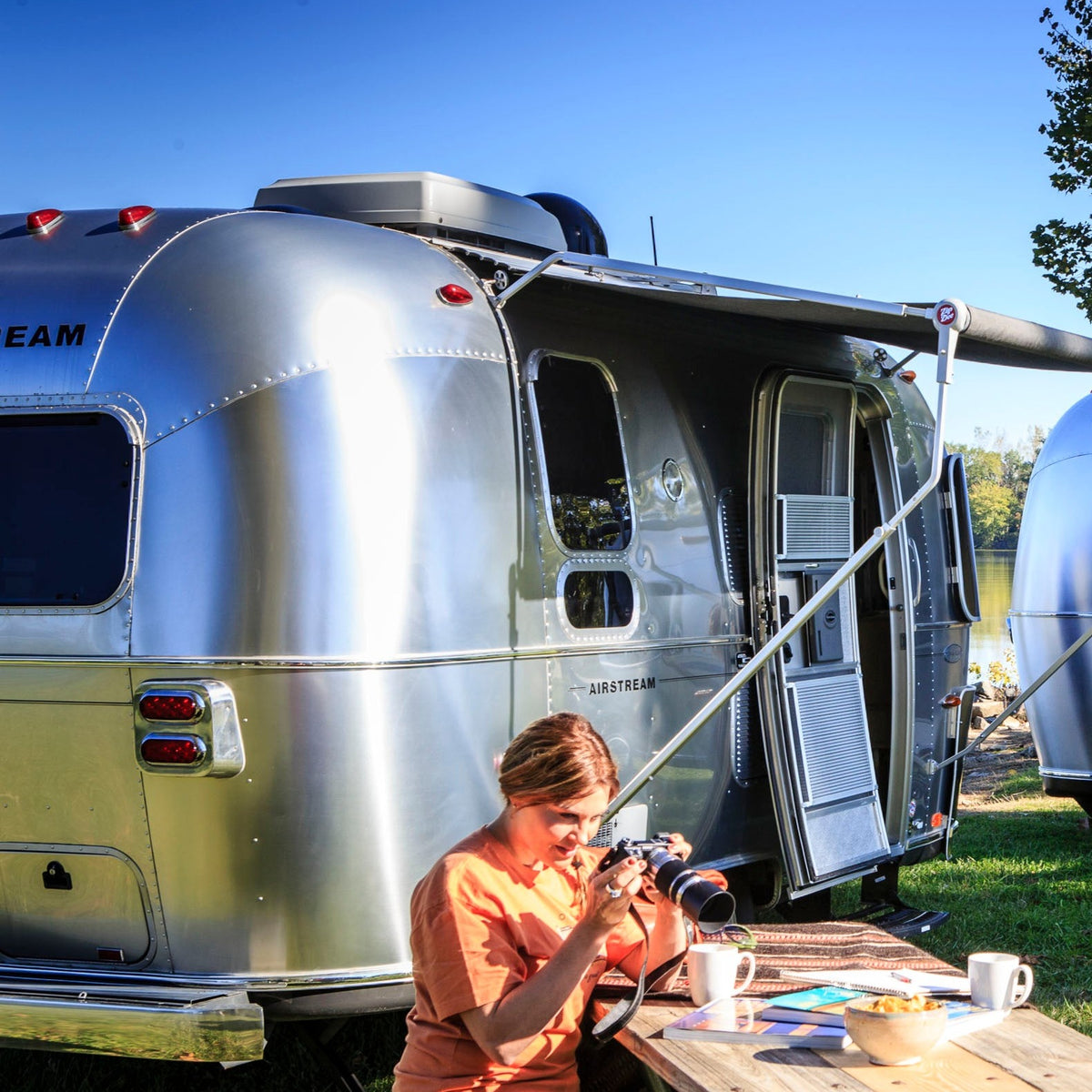 A person sits at a picnic table beside a shiny silver Airstream trailer, examining a camera. The iconic Airstream Black Logo Decal (Part #386150, 386151) gleams on the trailer’s side, complementing the mugs, notebook, and other items scattered across the table. Trees and a blue sky create a peaceful backdrop for this scene.