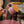 A close-up of a person's hands using a metal scraper to remove excess pink paint from the handle of a pink ceramic mug, which is balanced on its orange base over a pink paint bucket on a workshop table.