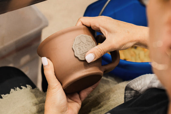 A person holds a handmade terracotta clay mug with a carved 'STOCKTON' logo on the side. The mug is shown in a close-up, lifestyle setting with hands holding it and a blue bag in the background.