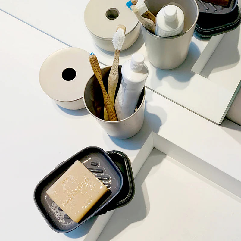 A Bamboo Travel Soap Box by Ekobo rests in a dish, while two toothbrushes and a toothpaste tube sit in a nearby cup; all reflected on a white bathroom countertop.