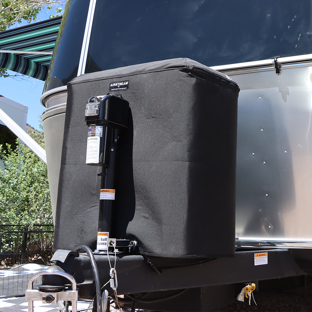A close-up of the Airstream Water Resistant Propane Tank Cover in black, protecting a propane tank on the front hitch of a silver trailer, with greenery and an awning visible in the background.