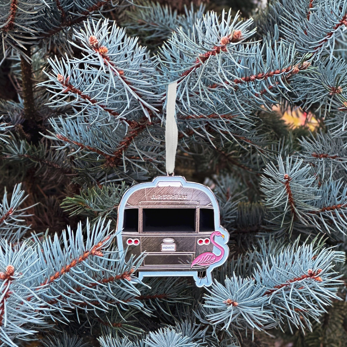 An Airstream Flamingo Travel Trailer Flat Ornament, featuring a classic silver camper and a pink flamingo, hangs from a blue spruce tree branch by a white ribbon, making it the perfect gift to celebrate the silver season.