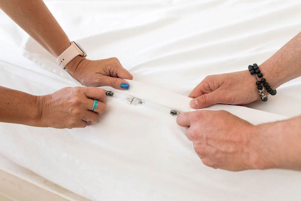 Two people zip together a white bedsheet—specifically, the Airstream RV Fit Beddy’s ZipBase Sheets for Classic Travel Trailers—each using both hands. One is wearing a smartwatch, a ring, and has blue nail polish; the other sports black beaded bracelets. The focus is on their hands and the sheet, perfectly highlighting how these sheets simplify bed-making in your Airstream or RV.